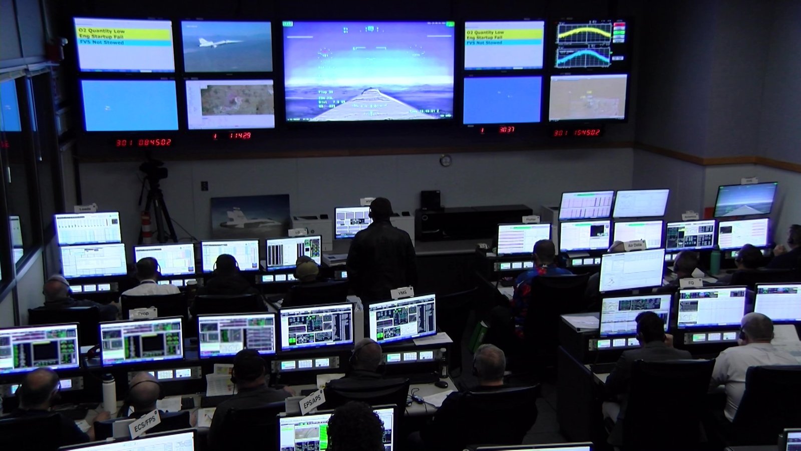 One large central screen, surrounded by four smaller screens on each side, spans the front wall of a control room displaying video and research flight data. Rows of engineers wearing headsets monitor real-time data on their computer screens.