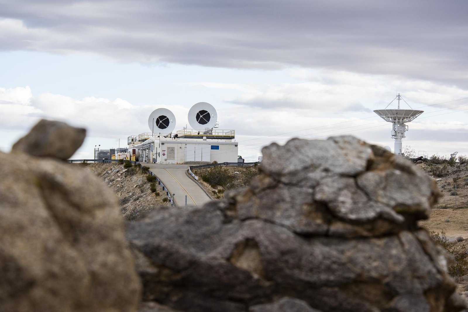 A large antenna points to the sky, while two large radar dishes are attached to the roof of a building to the left. Rocky terrain stretches across the foreground. 
