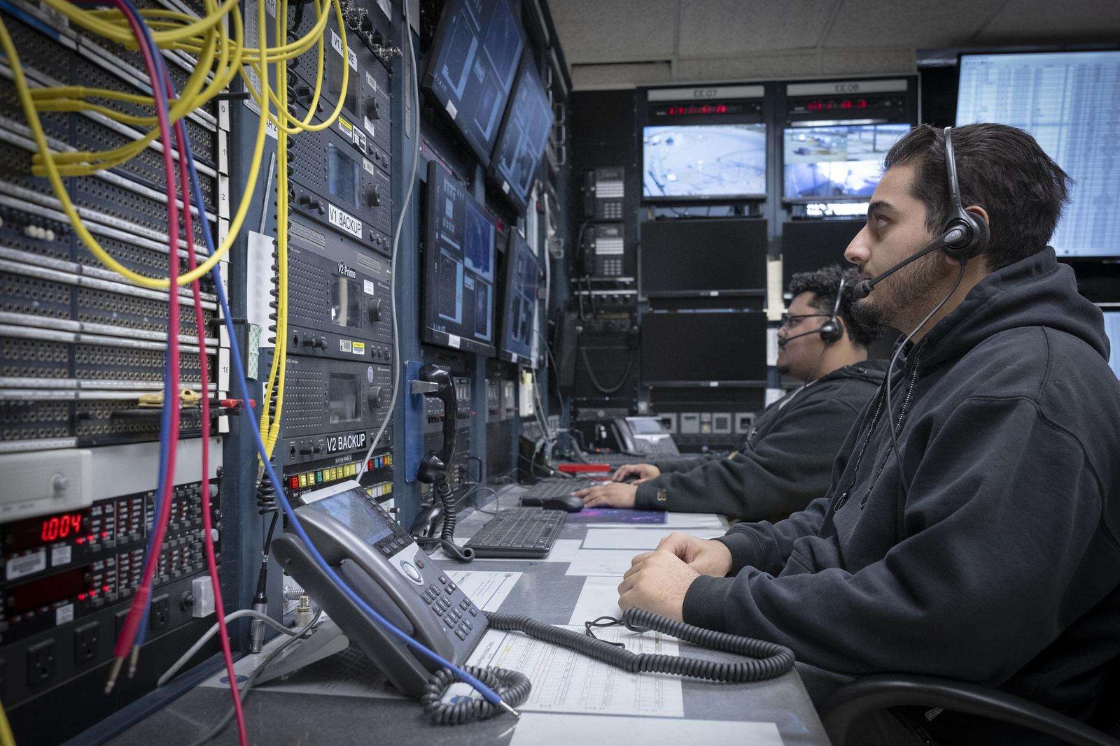 Two men wearing headsets sit in front of a wall of screens and instrumentation.
