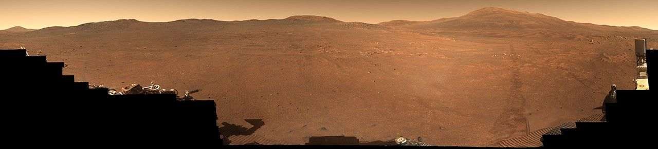 A rock-strewn, brownish-red Martian plain is bordered by a series of ridges and hilltops on the horizon, with rover tracks leading from them to the foreground on the right side of the image, where small portions of Perseverance rover are visible.