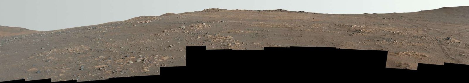 Undulating terrain of reddish-brown soil and loose rocks stretches toward a rugged horizon. The sky above is hazy and pale. The image’s bottom edge is a jagged black border, indicating the edges of the frames stitched together to create the mosaic.