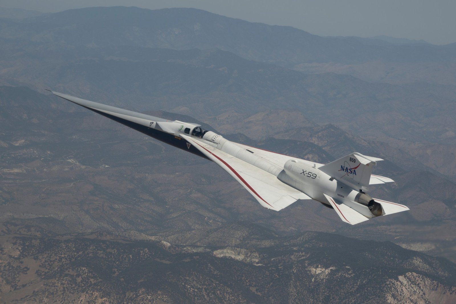 NASA’s X-59 quiet supersonic research aircraft flying above mountain ridges in California during a test flight on May 12, 2026. The aircraft is viewed from above, showing its elongated nose, swept wings, and white fuselage against layered mountains and valleys below.