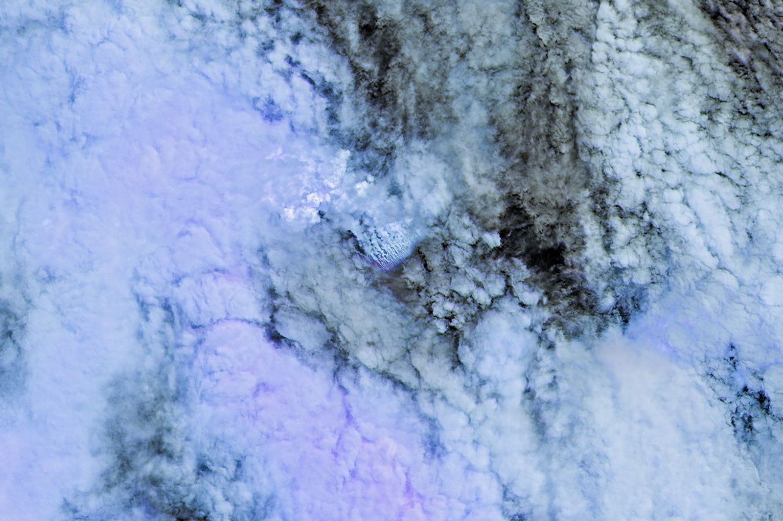 Only small patches of the ocean are visible through puffy, white volcanic plumes streaming from the center of the scene. Clouds not directly related to the eruption are visible throughout much of the image. A false-color inset box shows the infrared signature of the eruption as a series of red dots near the volcanic plumes.
