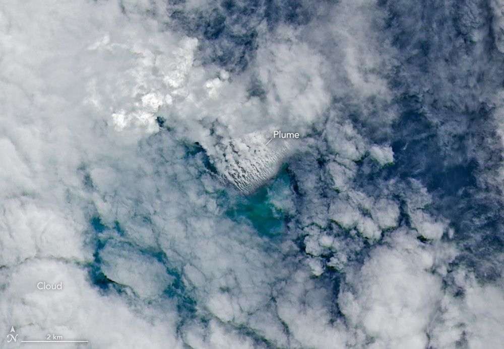 Only small patches of the ocean are visible through puffy, white volcanic plumes streaming from the center of the scene. Clouds not directly related to the eruption are visible throughout much of the image. A false-color inset box shows the infrared signature of the eruption as a series of red dots near the volcanic plumes.