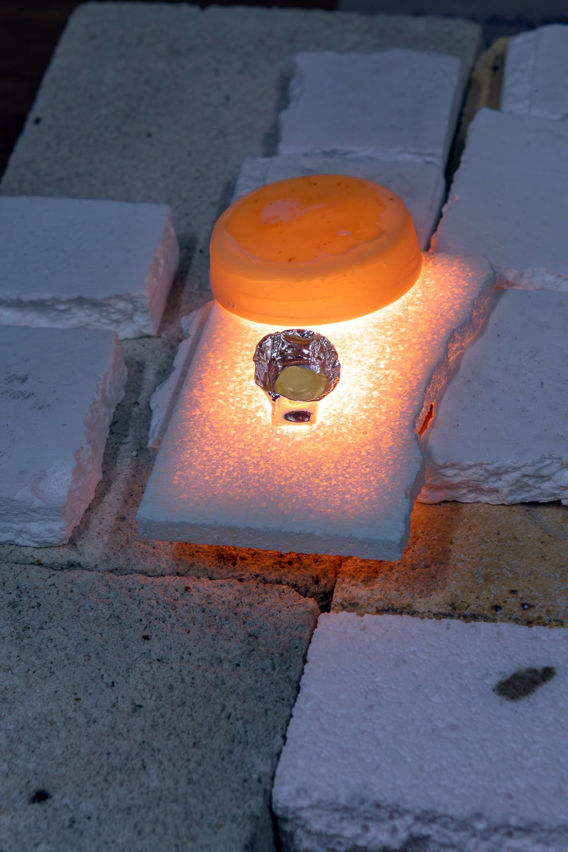 A melted tan material sits inside of a small silver-colored container. A larger glowing orange dome is propped up behind the container. Both items are laying on top of a white and grey surface with a brick-like texture.