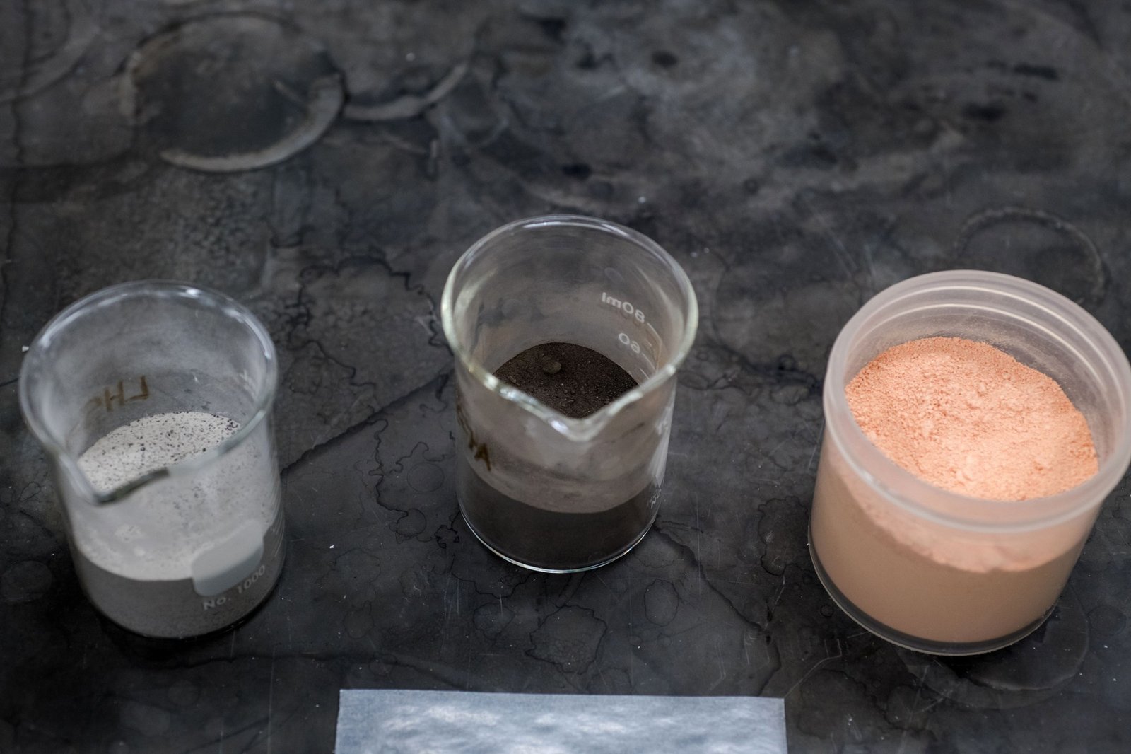 From left to right: a top-down view of small beaker containing light grey powder, a small beaker containing black powder, and a jar containing bright pink powder. They all sit on top of a greyish-black counter.