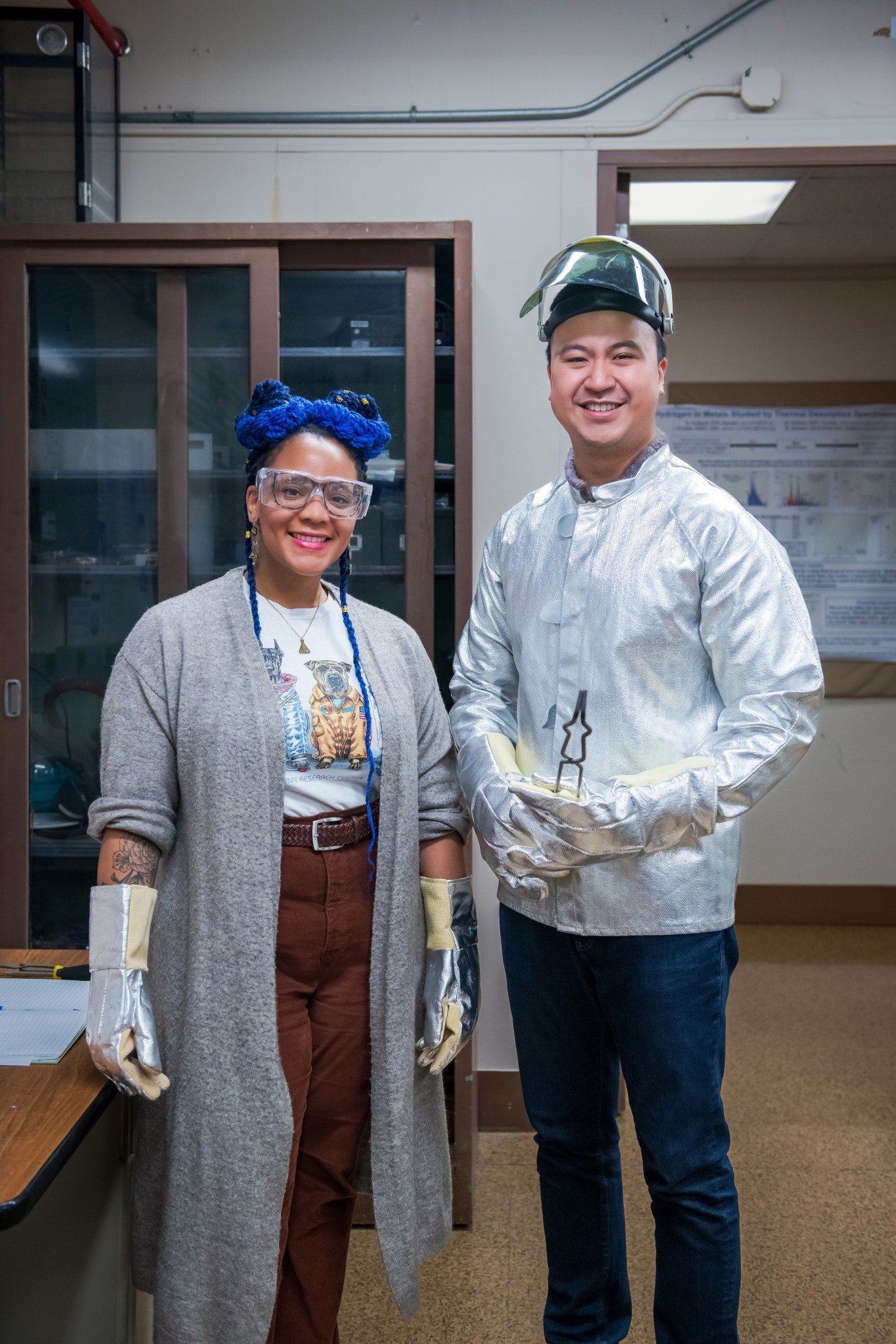 Two researchers pose for a portrait inside a laboratory at NASA Glenn. The researcher on the left is wearing a long grey sweater, silver protective gloves, and safety glasses. The researcher on the right is wearing a silver protective coat, helmet, and gloves and is holding large tongs. 