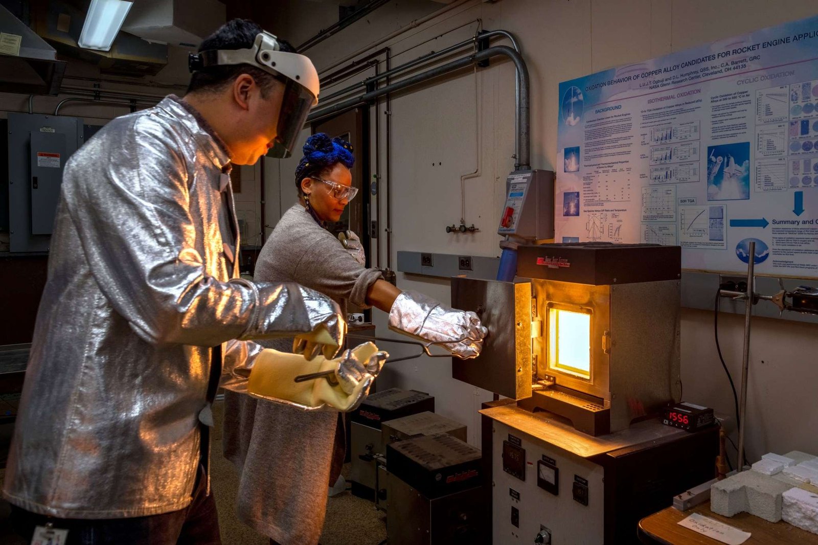 Two researchers wearing shiny, silver-colored protective gear including thick gloves, coats, and eyewear stand beside a furnace inside a laboratory as they prepare to remove a sample of a new material. The researcher on the left holds large tongs and reaches toward the furnace as the researcher on the right holds open the furnace door. The inside of the furnace is glowing bright white-orange.