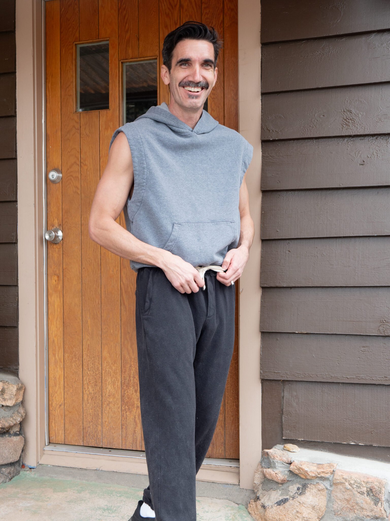 A man with a mustache stands outside a wooden door, wearing a sleeveless gray hoodie, black sweatpants, and black sneakers, holding the waistband of his pants.