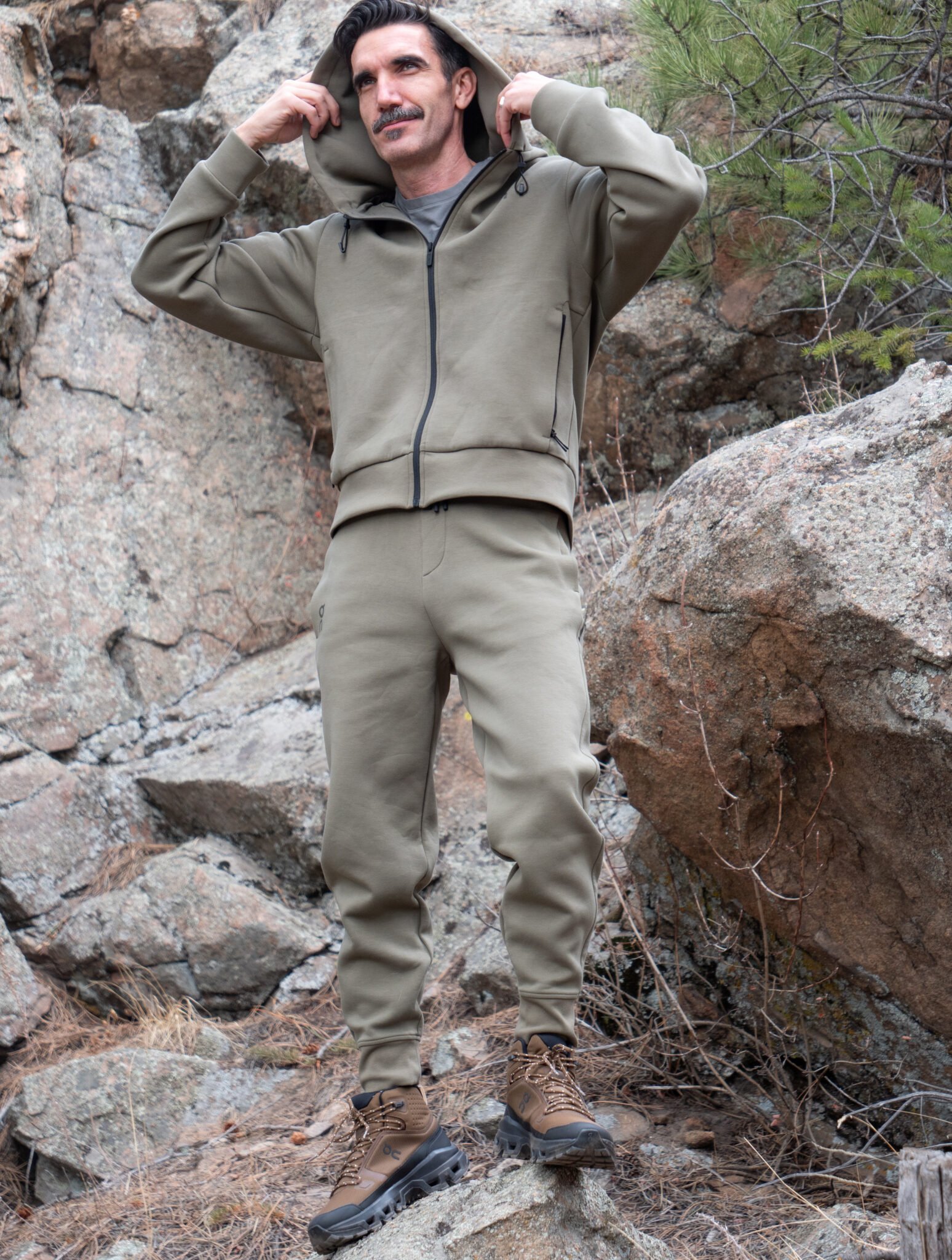 A man wearing a khaki hoodie and joggers stands on rocky terrain outdoors, holding the hood of his jacket with both hands.