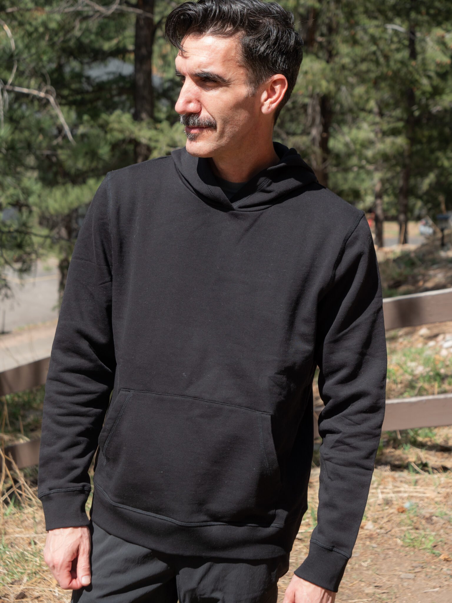 Man with a mustache wearing a black hoodie stands outdoors in a wooded area, looking to the side.