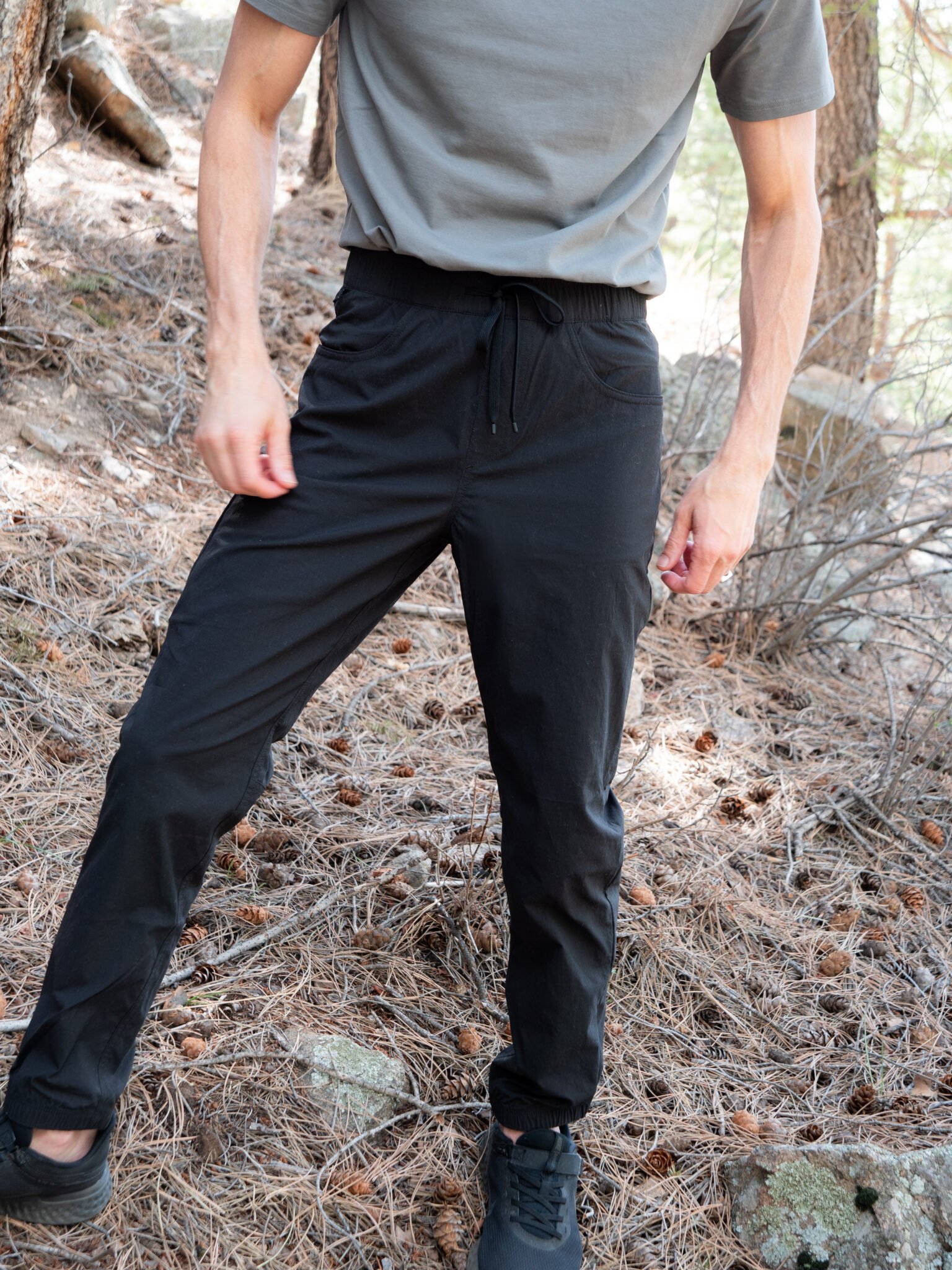 A person standing outdoors on a forest floor, wearing a gray t-shirt, black pants with a drawstring, and black shoes. Their face is not visible.