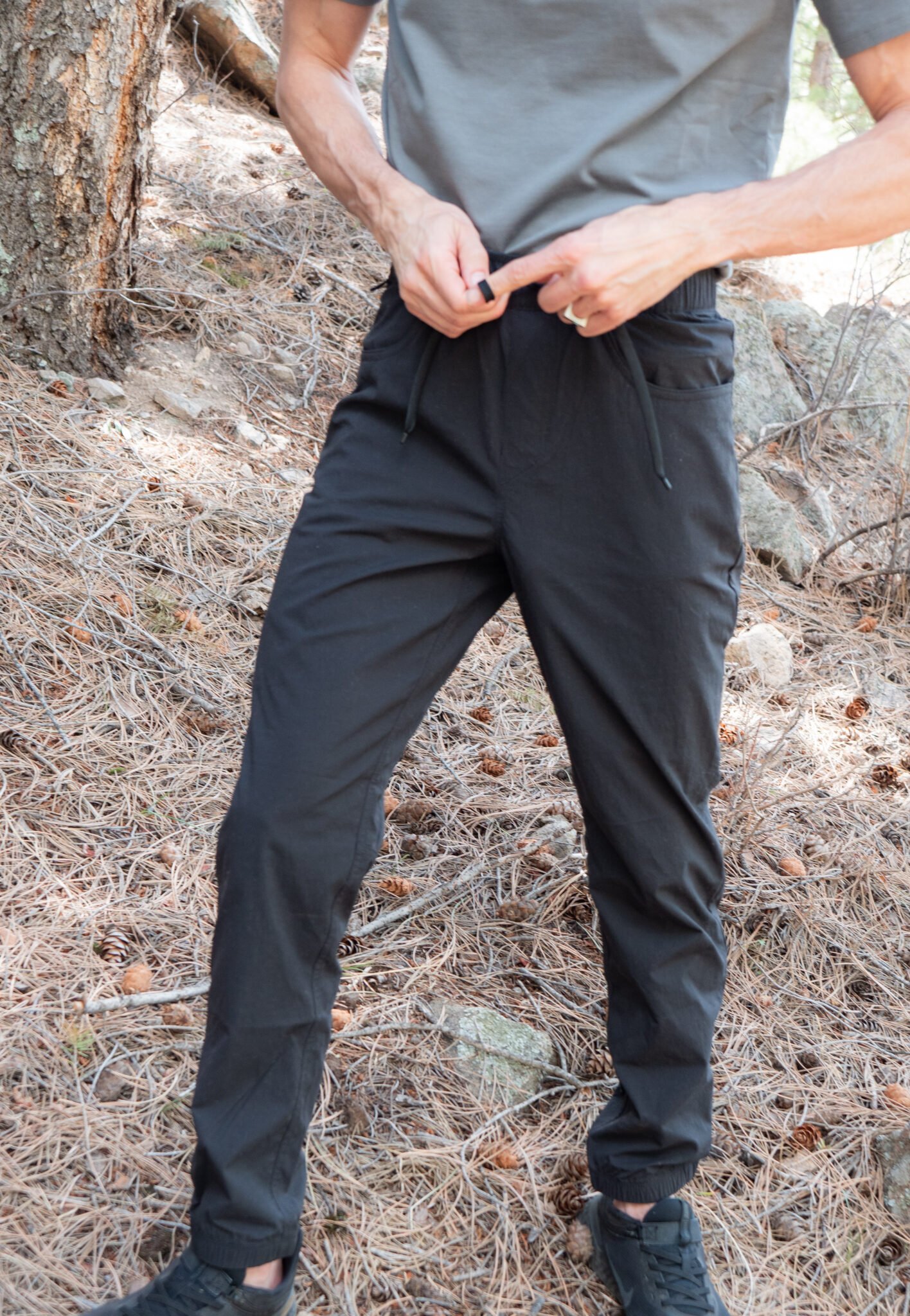 A person in a gray shirt and black jogger pants stands outdoors on pine-covered ground, adjusting the waistband of their pants.
