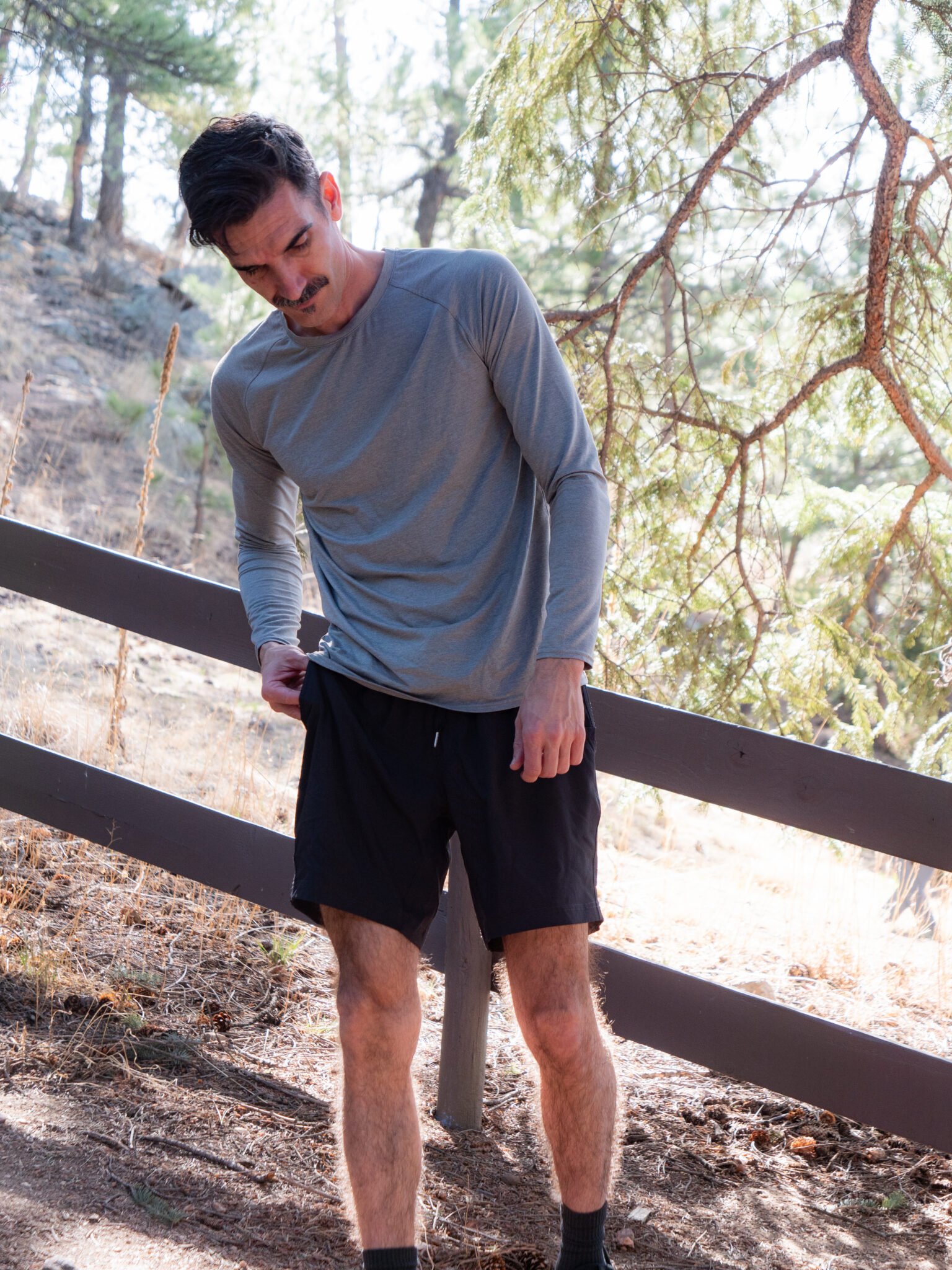 A man in a gray long-sleeve shirt and black shorts stands outdoors on a dirt path near a wooden fence and pine trees, looking down and adjusting his shirt.