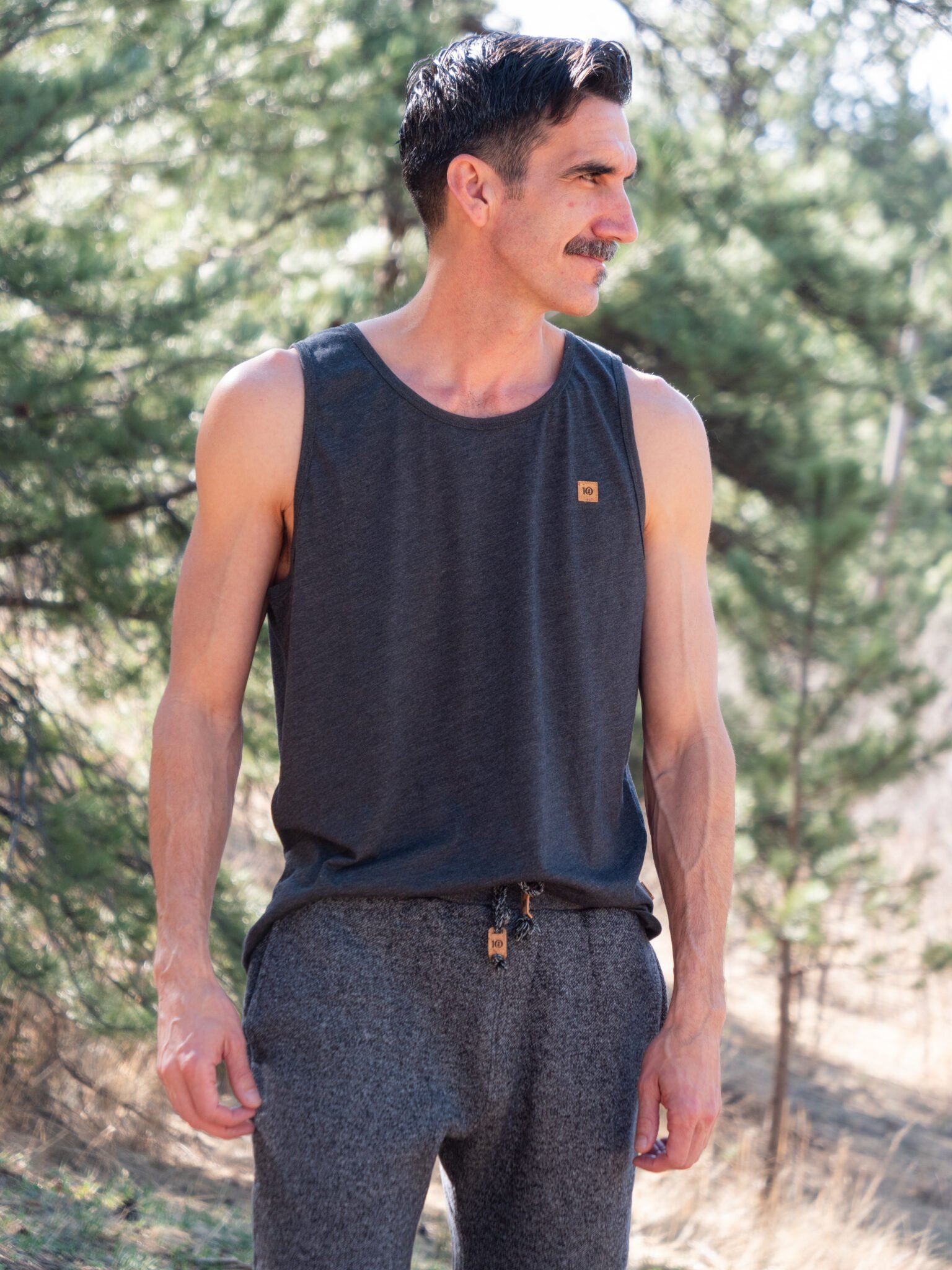 A man with dark hair and a mustache stands outdoors wearing a dark sleeveless shirt and gray pants, with trees in the background.