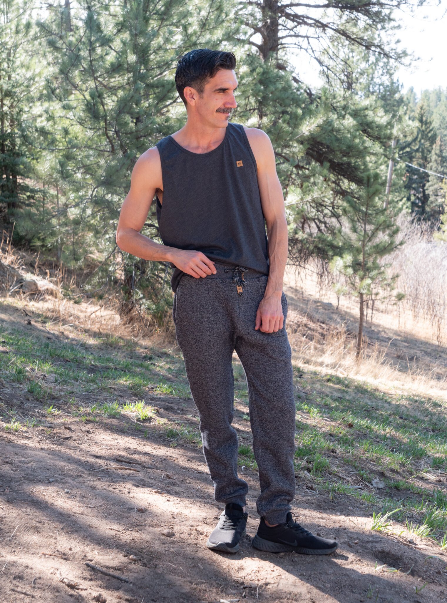 A man stands outdoors on a dirt path surrounded by trees, wearing a sleeveless dark shirt, gray jogger pants, and black shoes.
