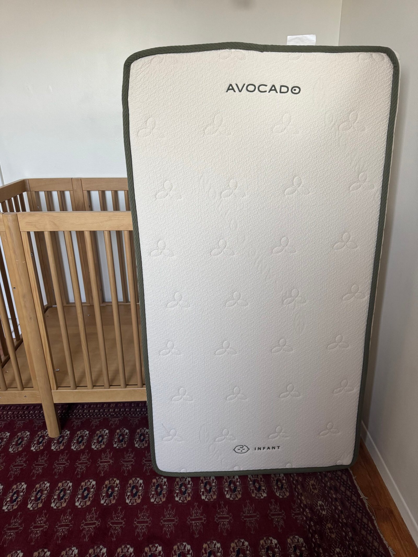 A crib mattress labeled "Avocado" stands upright next to a wooden crib on a red patterned rug in a room with white walls.