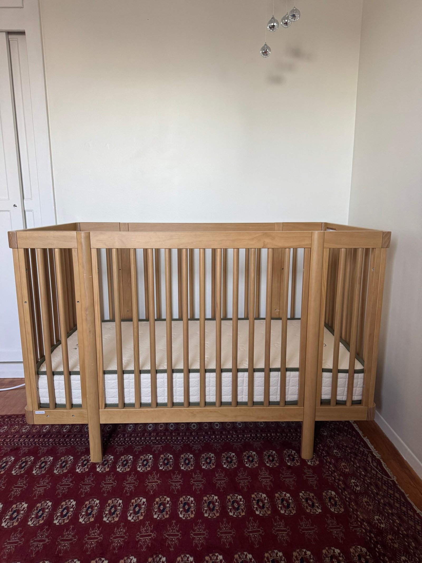 A wooden baby crib with a mattress stands on a patterned red rug against a plain white wall.