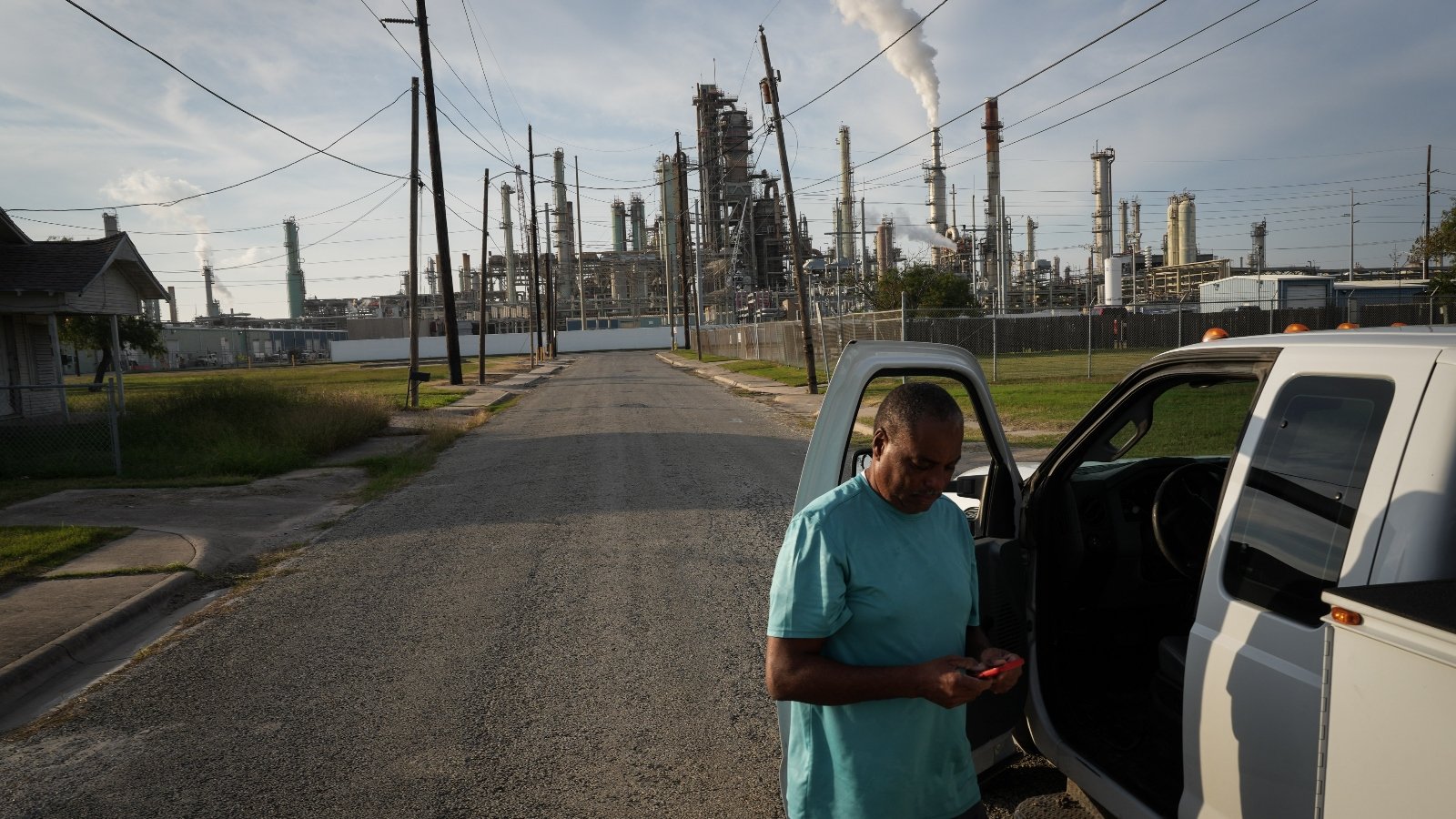 A man stands looking at his phone next to the open door of a truck, with a power plant spewing smoke in the background