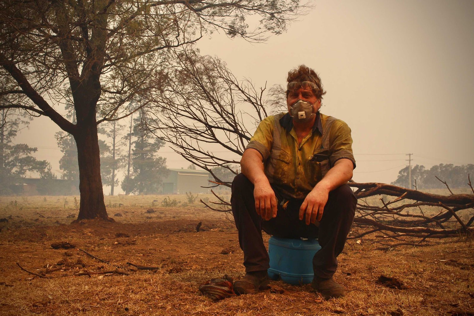 A man wearing a face mask is sitting down. Smoke and burned vegetation are in the background.