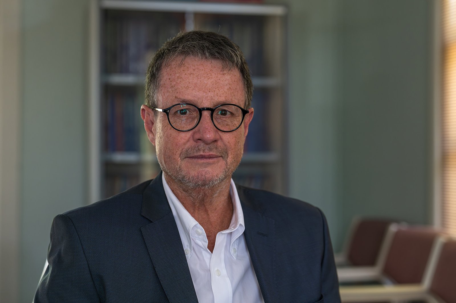 A man wearing glasses and a blazer sits for a portrait.