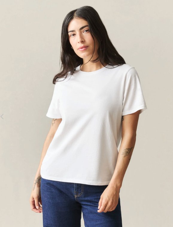 Woman standing against a plain background, wearing a white short-sleeve t-shirt and blue jeans, with visible tattoos on her forearms.