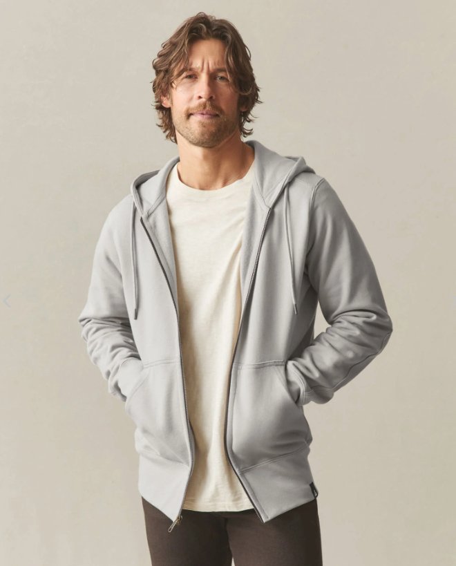 Man standing against a plain background, wearing a light gray zip-up hoodie over a cream-colored t-shirt with dark pants.