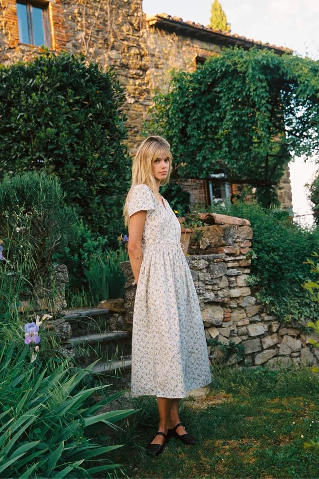 A woman in a light-colored floral dress stands outdoors near stone steps, surrounded by greenery and an old stone building in the background.