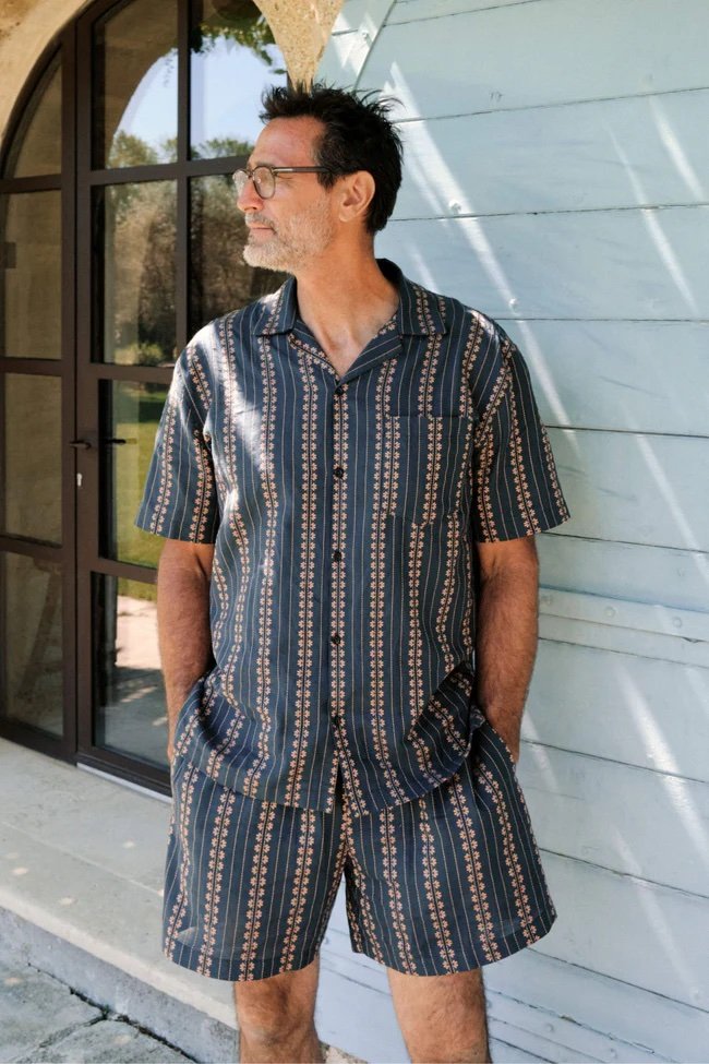 A man wearing a matching short-sleeve shirt and shorts with a patterned design stands outdoors by a wooden wall and arched window, looking to the side with his hands in his pockets.