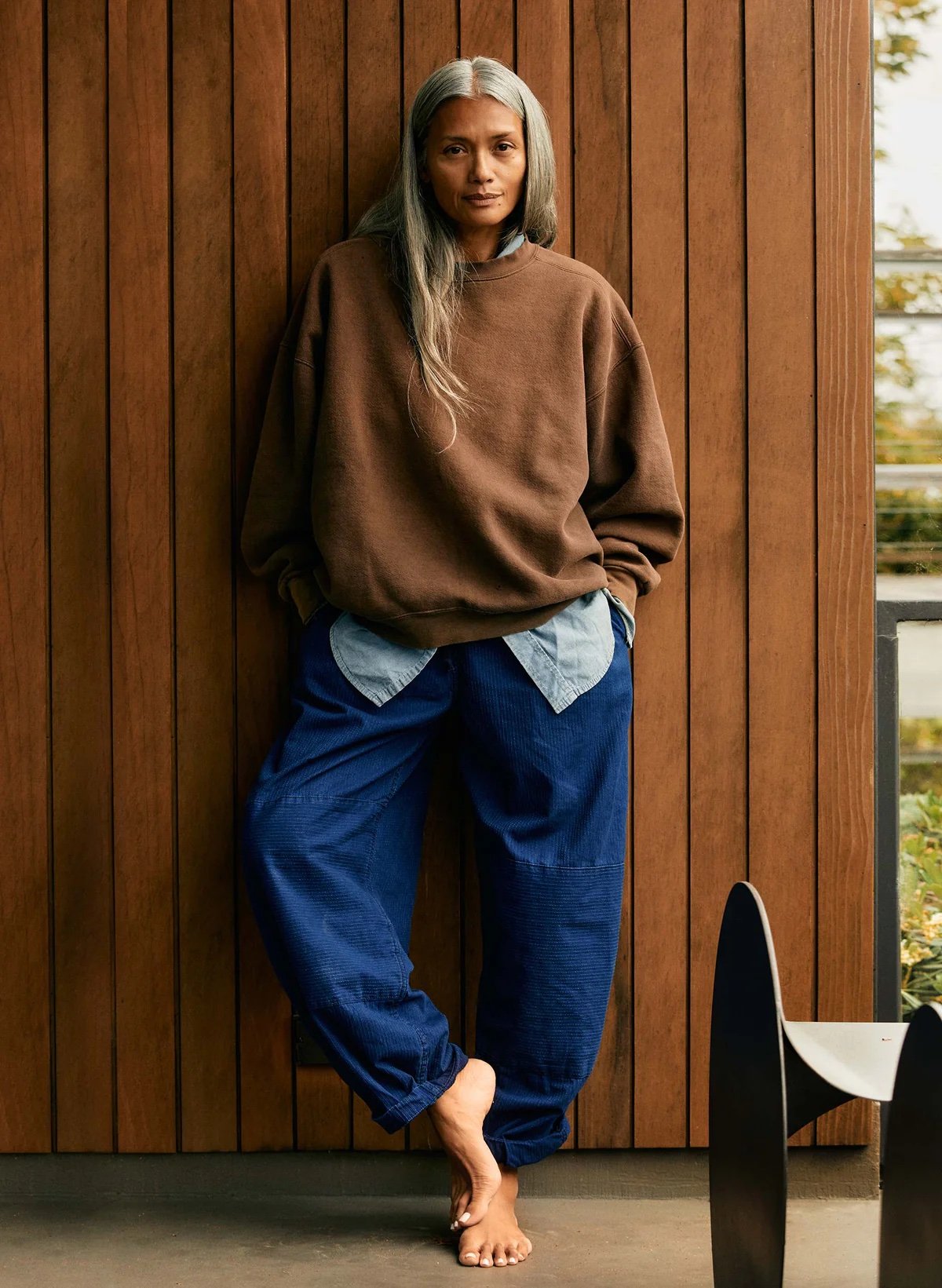 A woman with long gray hair stands barefoot against a wooden wall, wearing a brown sweatshirt over a blue shirt and loose blue pants.
