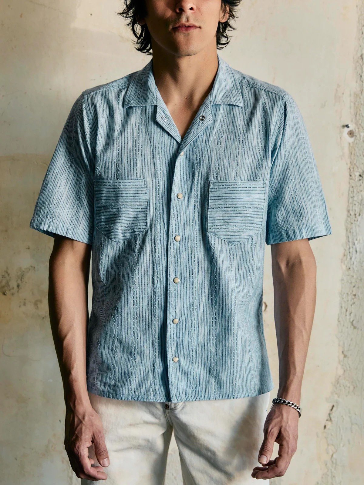A man wearing a light blue, short-sleeve, button-up shirt with two chest pockets, standing against a textured beige wall.