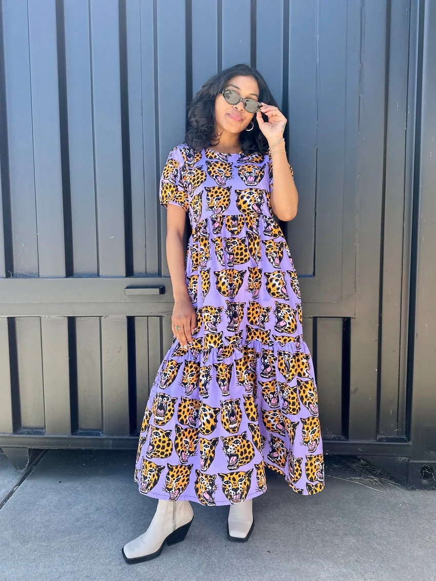 Woman stands in front of a dark gate wearing a long, purple dress with a yellow and black animal print, light-colored boots, and sunglasses.
