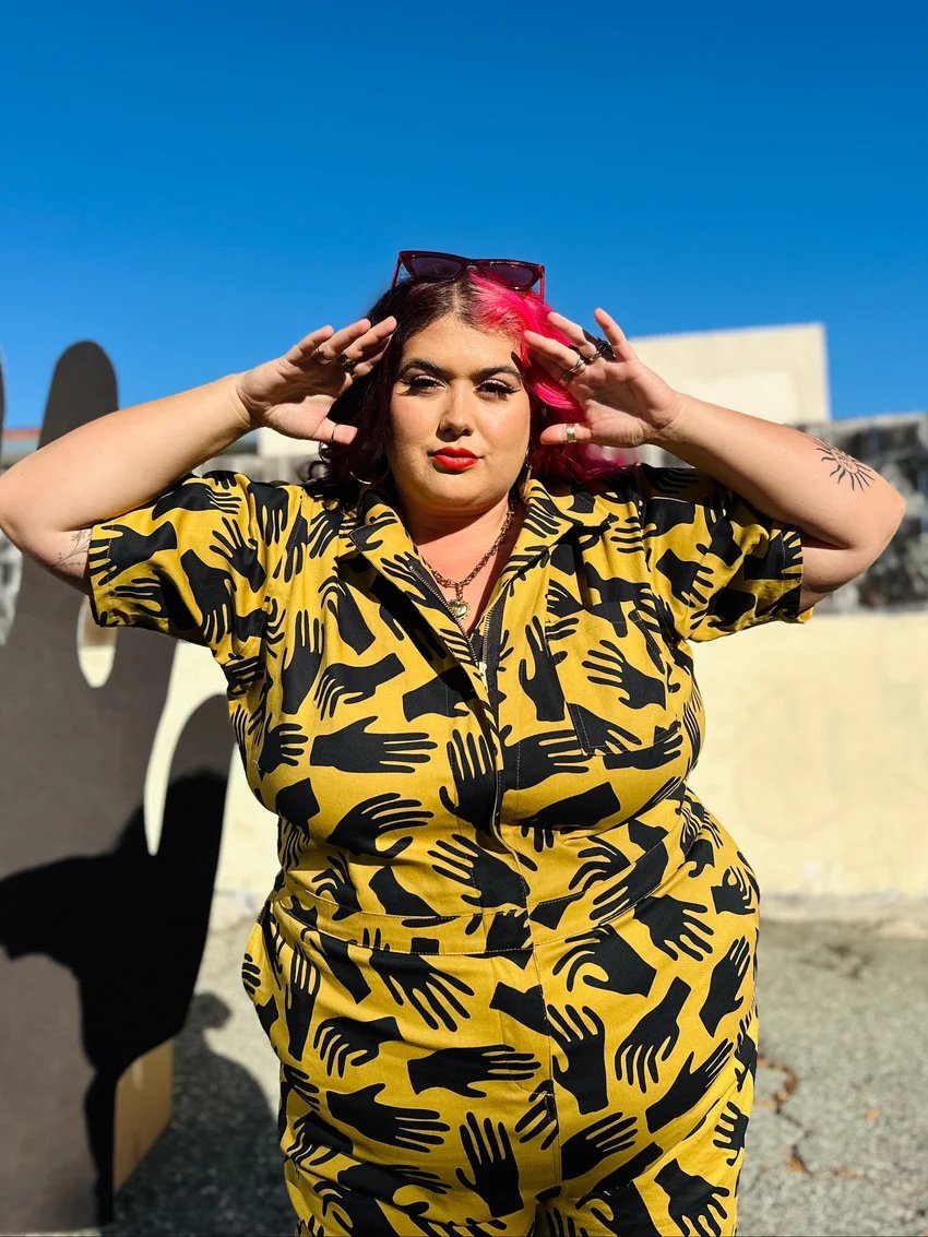 Person with pink hair wearing a yellow outfit with black handprints stands outdoors, holding their hands near their face with a neutral expression.