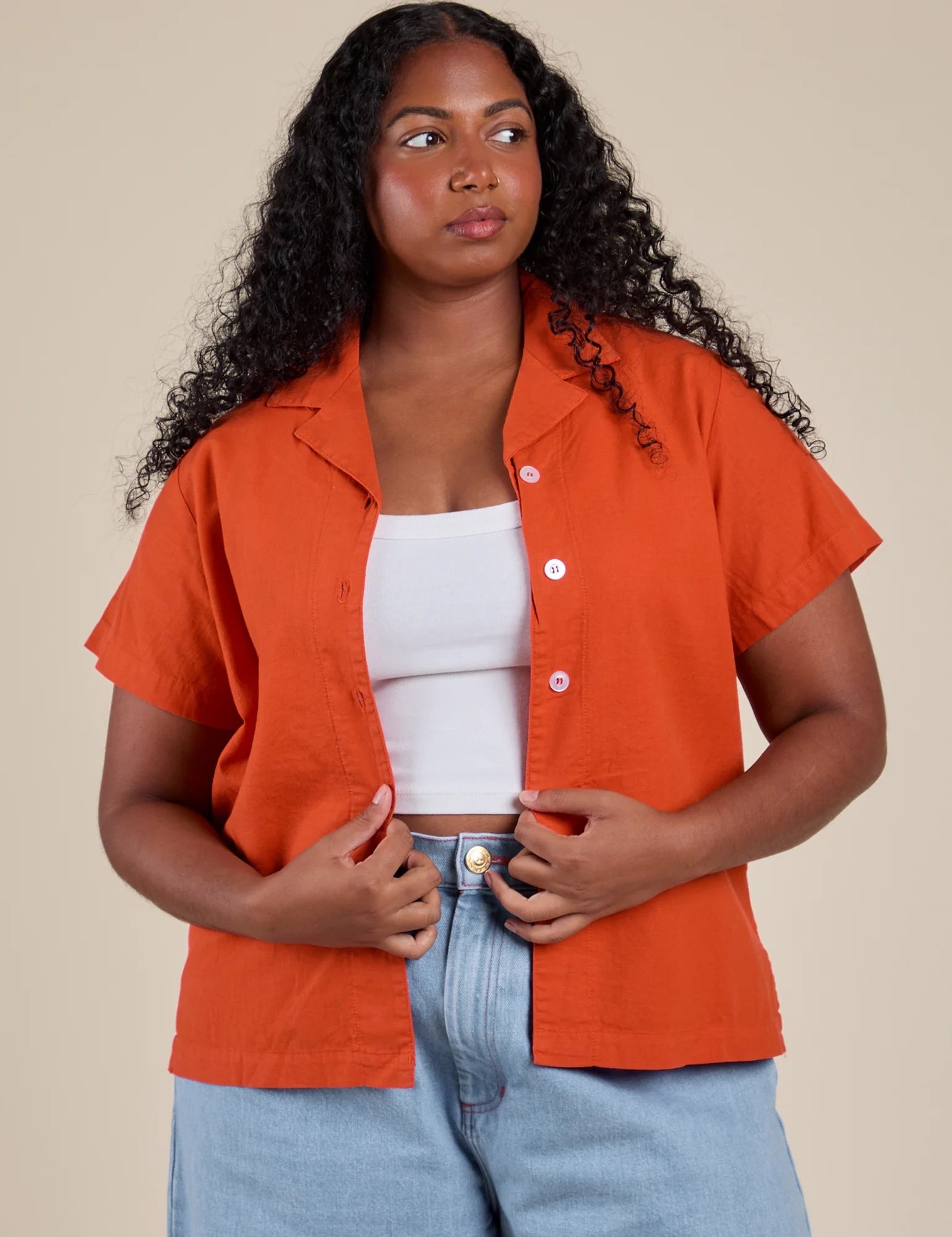 A person with long curly hair wears an orange short-sleeve button-up shirt over a white top and light blue jeans, standing against a beige background.