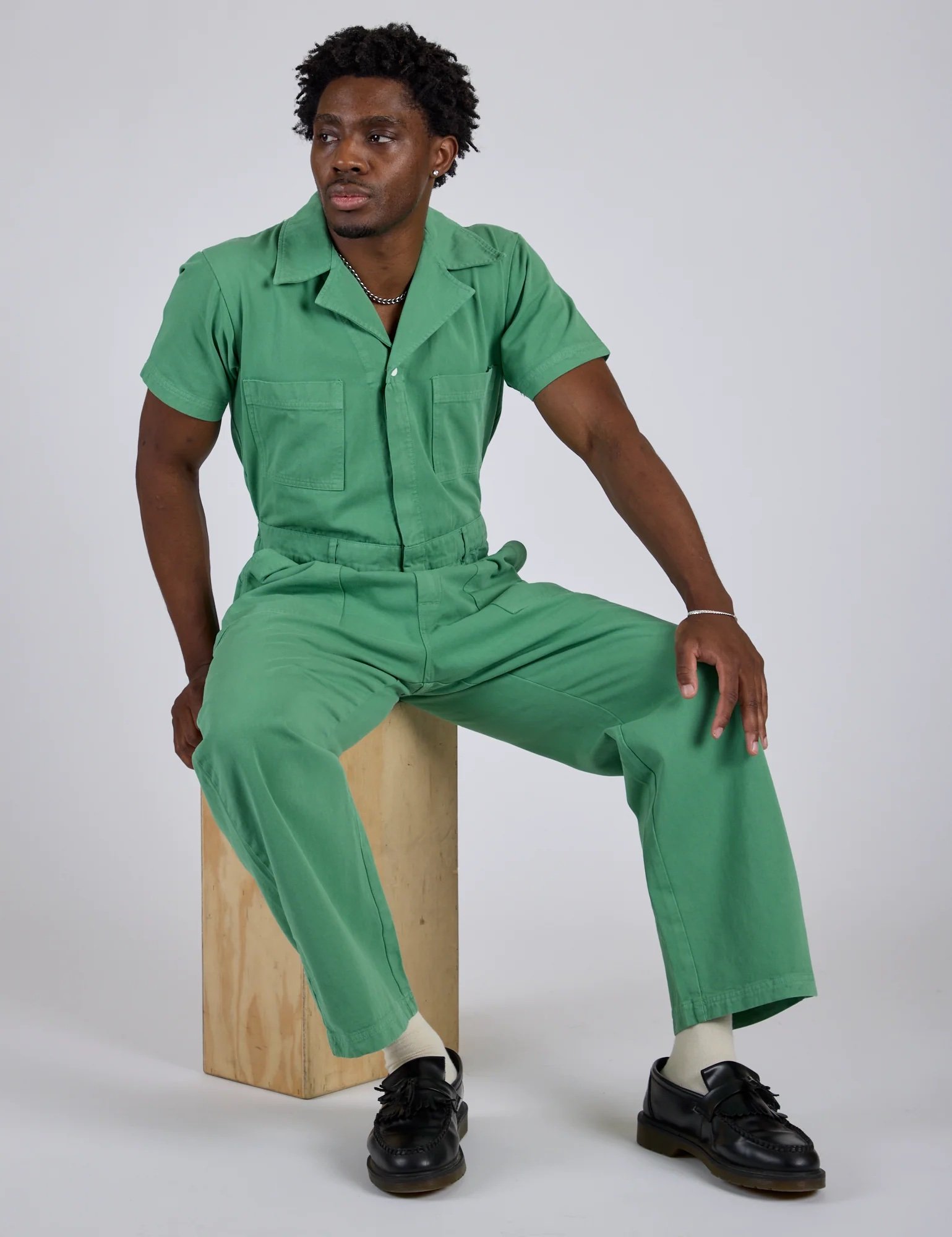 A person wearing a green short-sleeve jumpsuit and black shoes sits on a wooden box against a plain background.