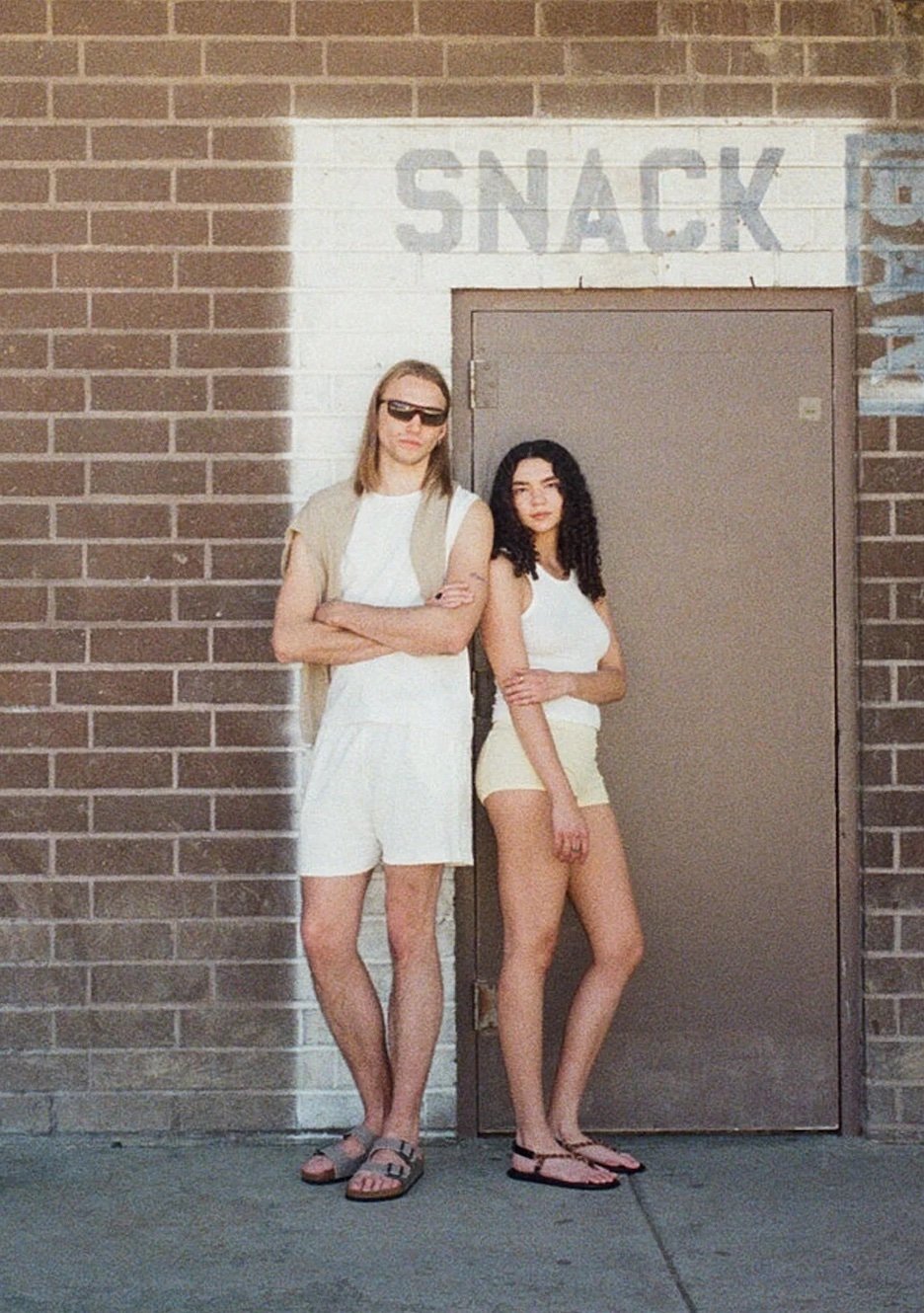 Two people stand side by side in front of a brick wall and a door with the faded word "SNACK" above it. Both wear casual summer clothing and sandals.