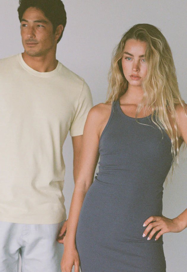 A man in a light yellow t-shirt stands next to a woman in a gray sleeveless dress; both look away from the camera with neutral expressions.