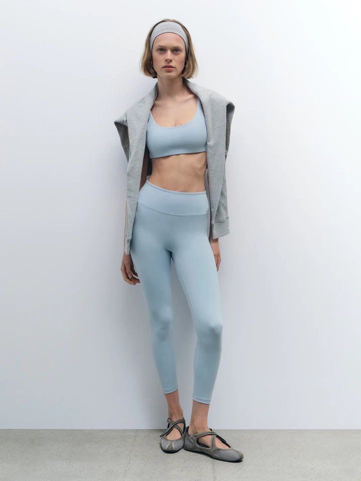 Woman standing against a plain wall wearing a light blue sports bra and leggings set, grey headband, grey sweatshirt draped over shoulders, and grey ballet flats.
