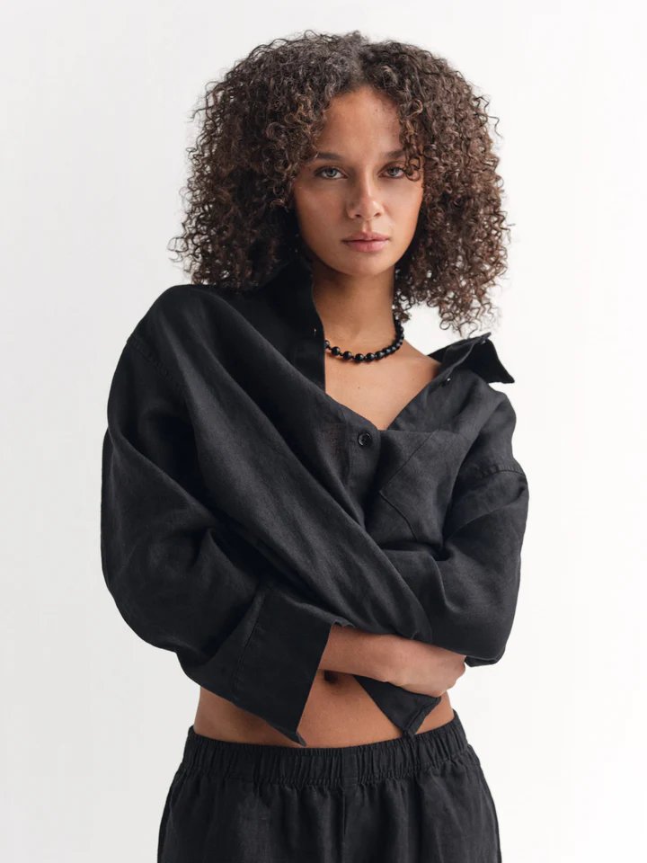 A woman with curly hair wears a loose black button-up shirt, black beaded necklace, and black pants, standing against a plain light background.