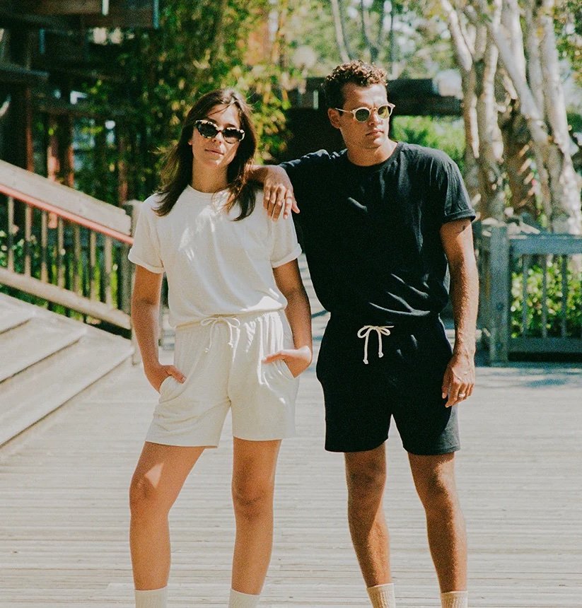 Two people stand outdoors wearing sunglasses and casual matching outfits; one in white, one in black. They both have hands in pockets, with one resting an arm on the other's shoulder.