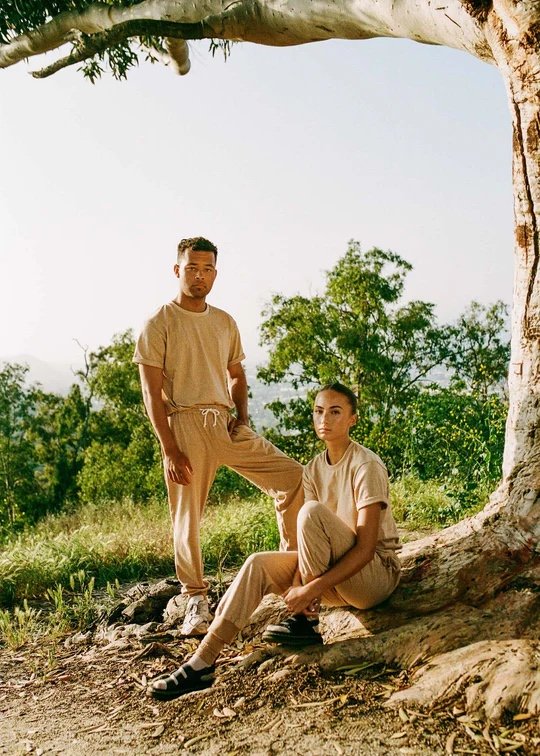 Two people in beige outfits pose by a tree in a natural outdoor setting with greenery and sunlight in the background.