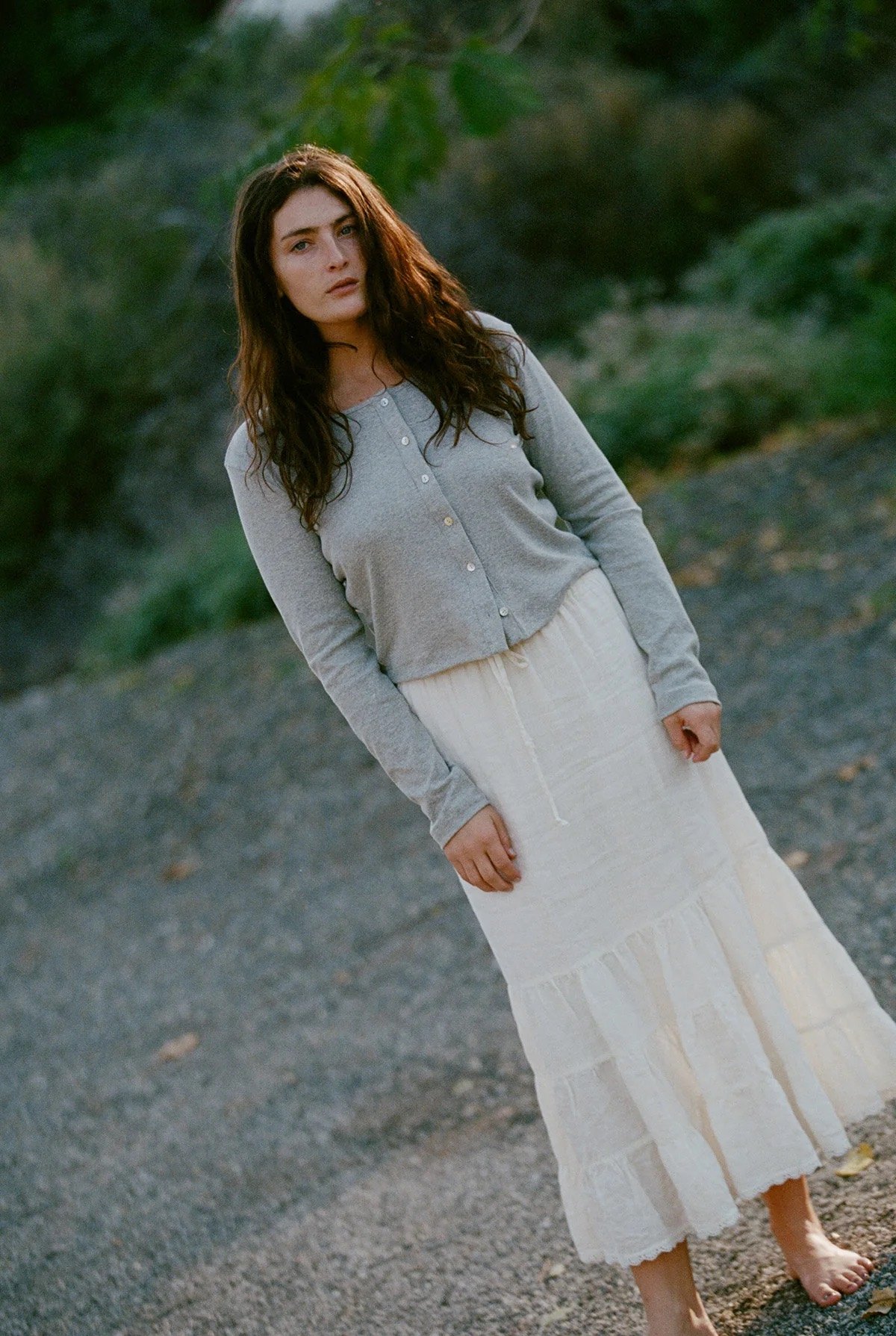 A woman with long brown hair stands outdoors on a gravel path, wearing a grey cardigan and a long white skirt, looking directly at the camera.