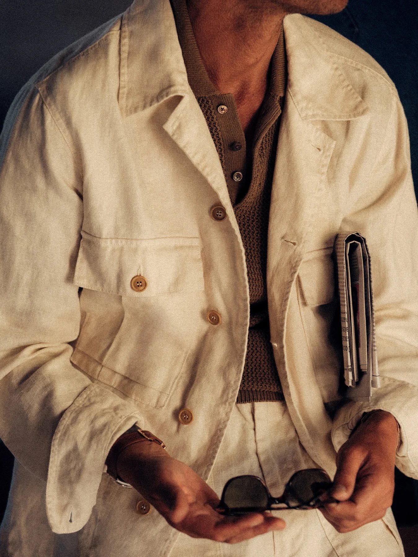 A person wearing a light-colored linen jacket and shirt holds sunglasses in one hand and a rolled magazine or document in the other, with the face partially out of frame.