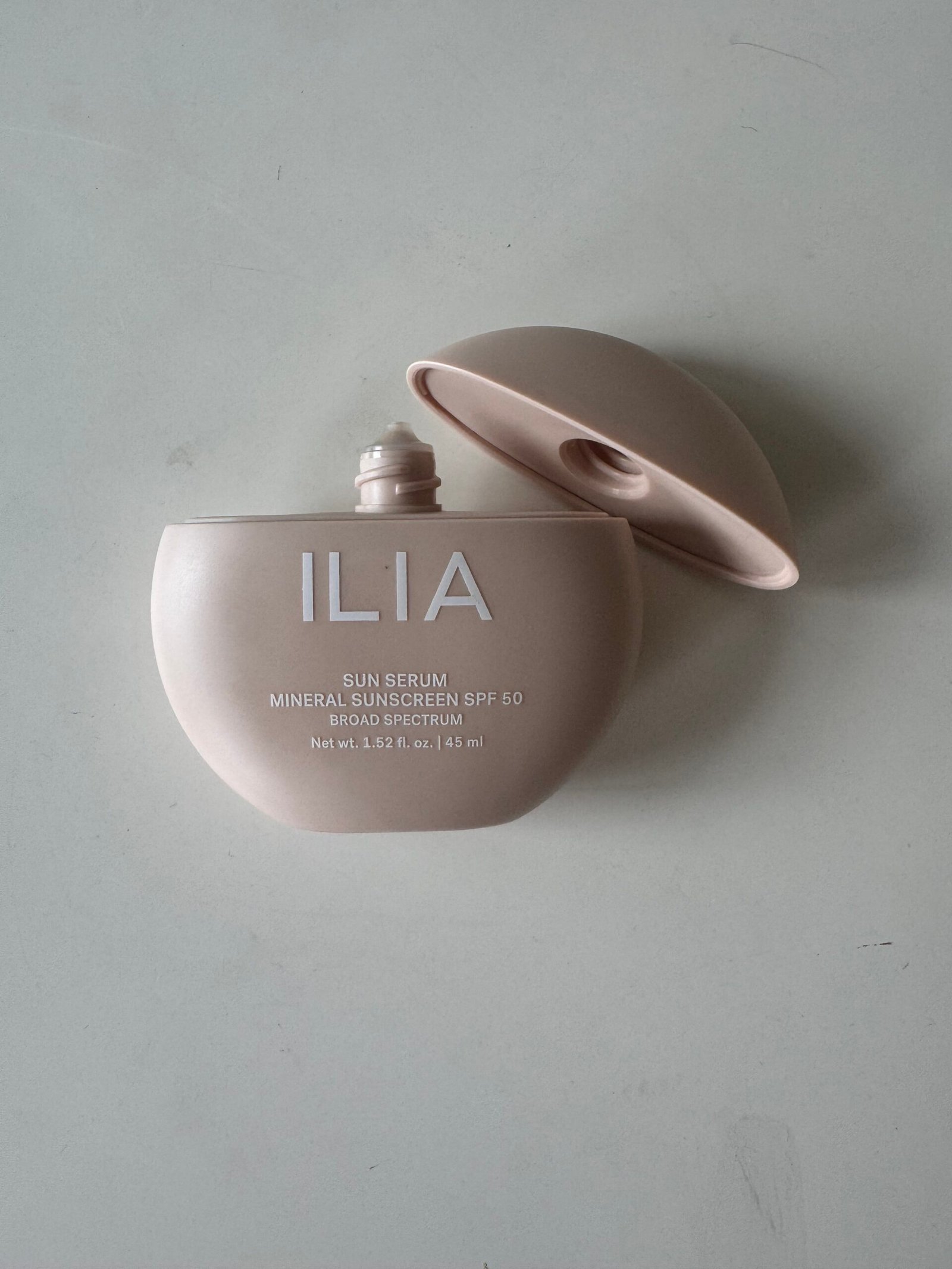 A bottle of ILIA Sun Serum Mineral Sunscreen SPF 50 with its lid off, placed on a plain light-colored surface.