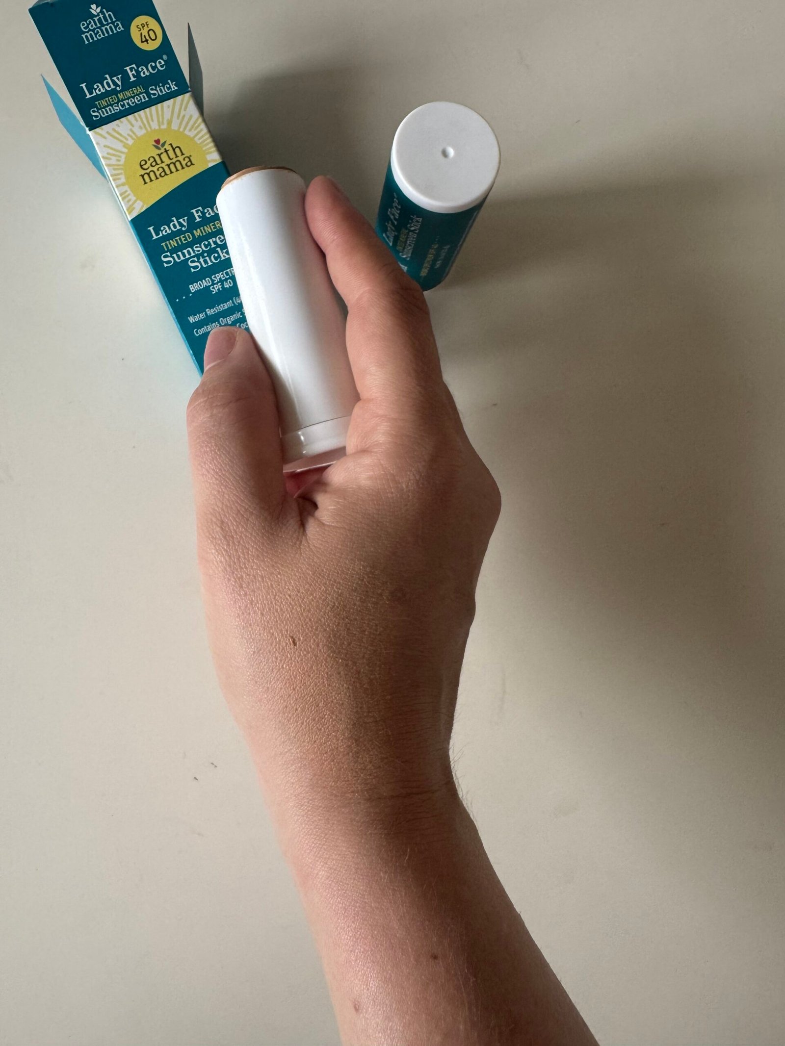 A hand holds a white tube labeled “Lady’s Face sunscreen stick” with its packaging and another stick on a light surface.