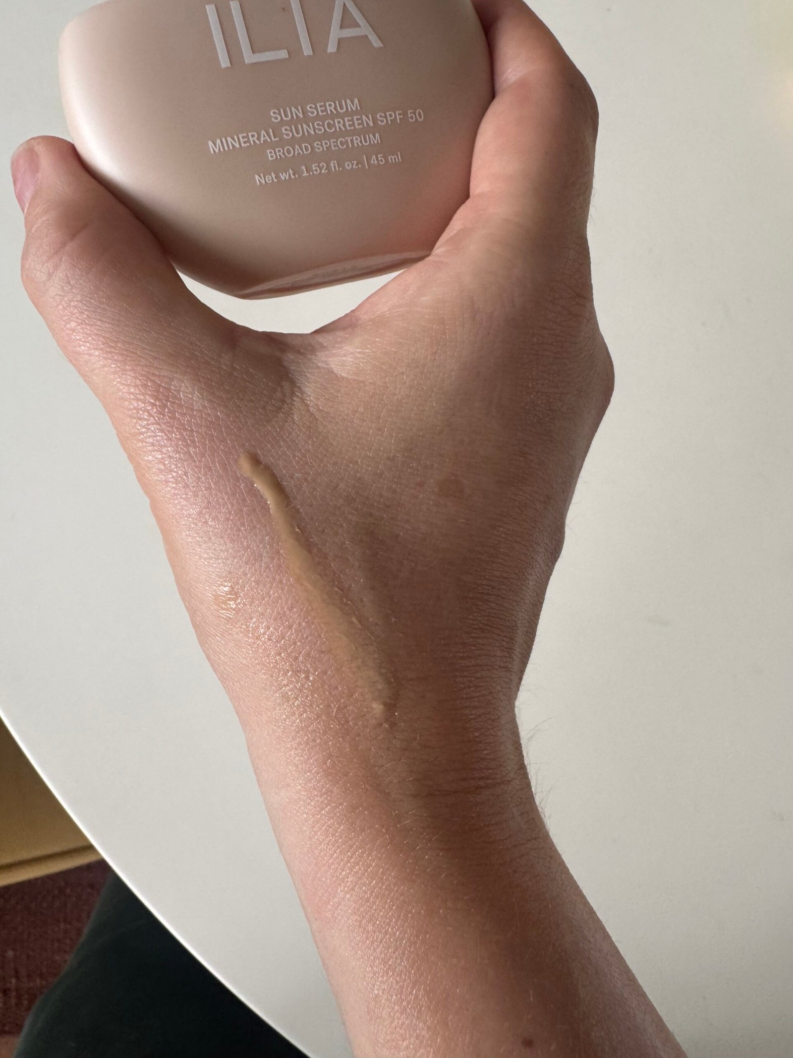 A hand with a streak of beige sunscreen on the back holds a bottle labeled "ILIA Sun Serum Mineral Sunscreen SPF 50.