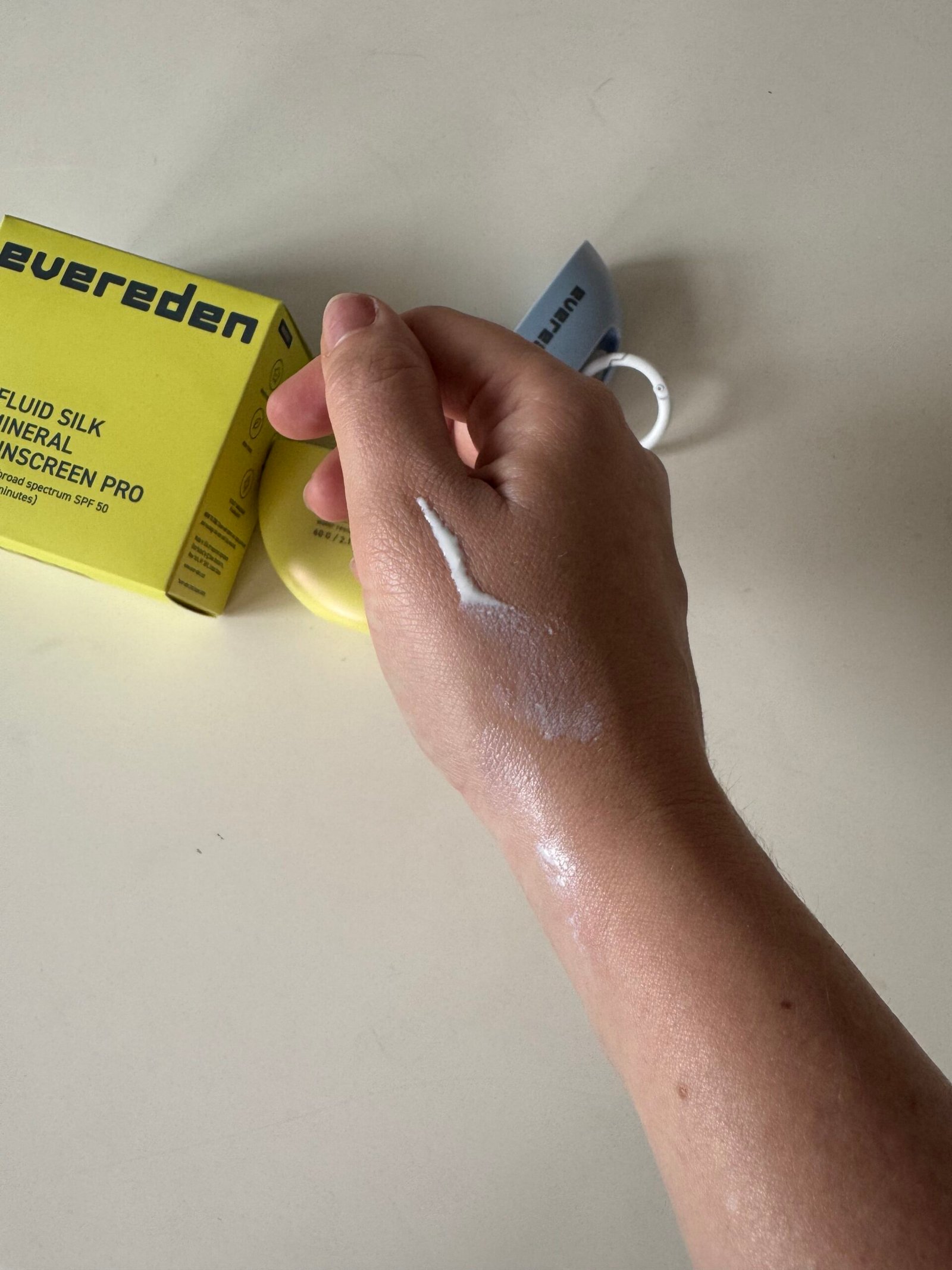 A hand with a streak of white sunscreen on the back is shown near a yellow Evereden sunscreen box and tube on a light surface.