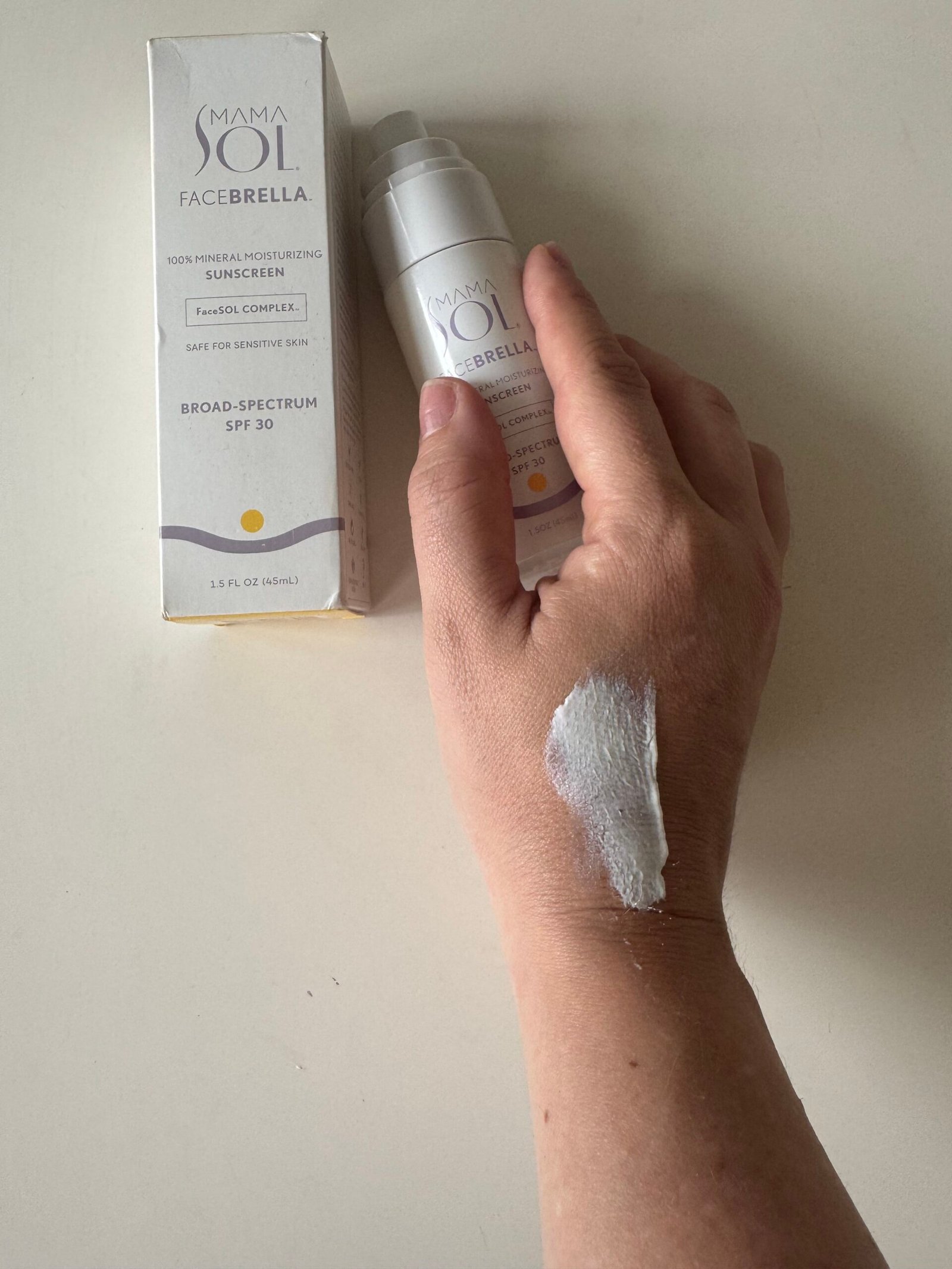 A hand with sunscreen applied on the back holds a tube of Mama Sol Facebrella SPF 30; the product box is placed beside the hand.