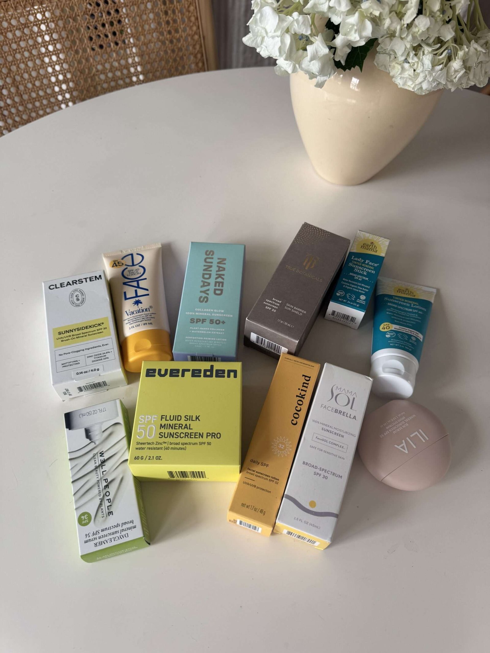 A variety of sunscreen and skincare product boxes arranged on a white table next to a cream vase with white flowers.