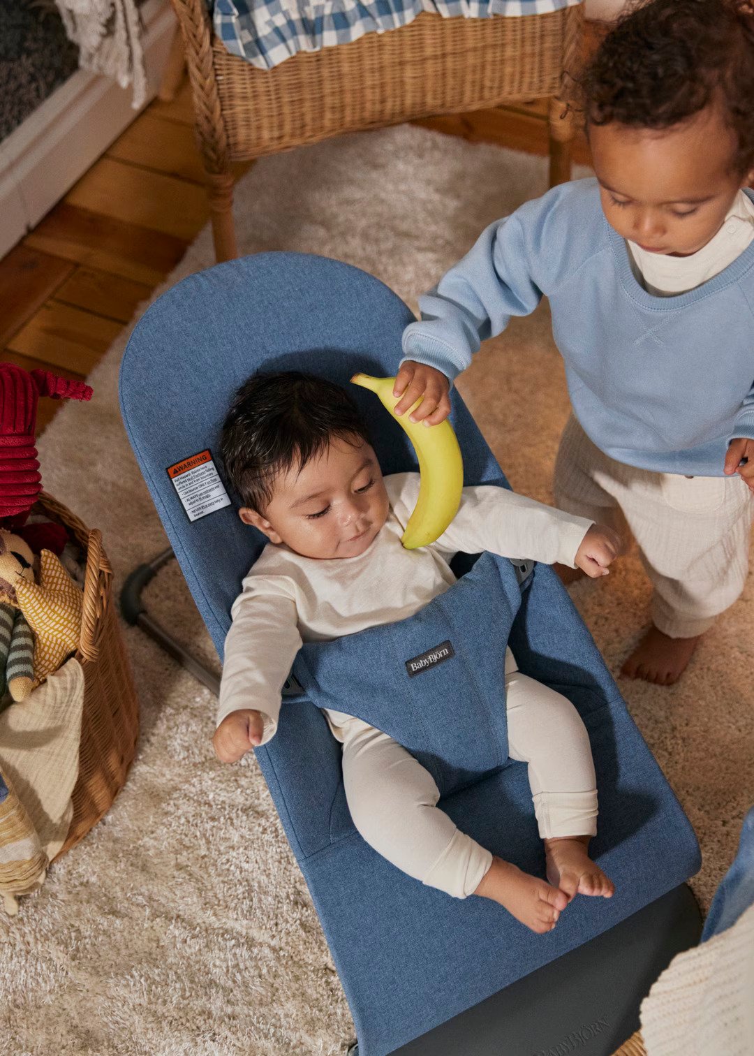 A baby sits in a blue bouncer while a toddler standing nearby holds a banana over the baby's head.